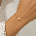 Hand wearing a gold bracelet with a pearl on a neutral background