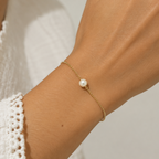 Hand wearing a gold bracelet with a pearl on a neutral background