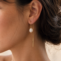 A close-up side profile of a woman wearing a delicate gold dangle earring with a white pearl-like drop, against a soft neutral background.